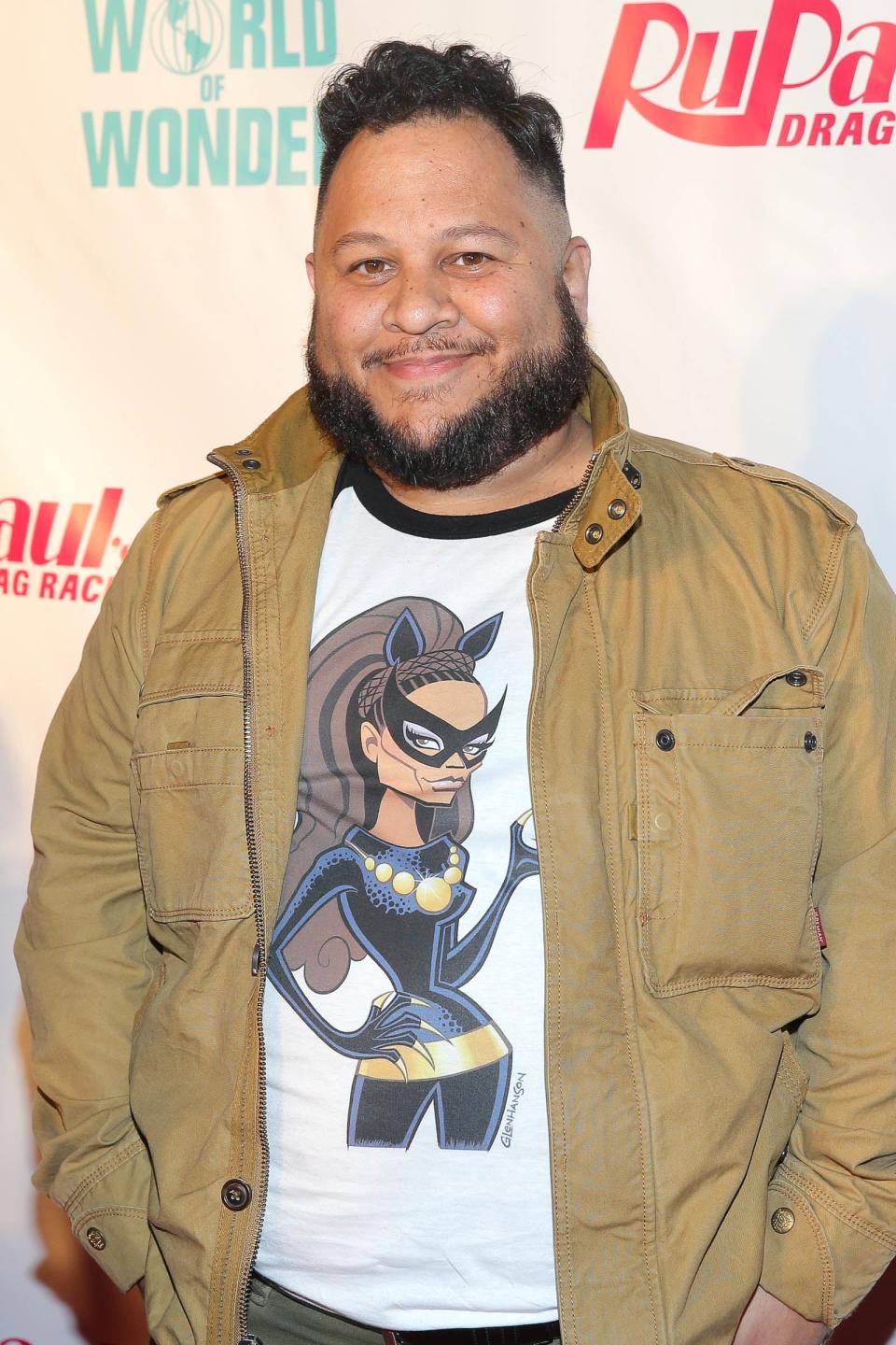 Person wearing a beige jacket over a graphic T-shirt with a superhero design, standing on a red carpet at the RuPaul's Drag Race event