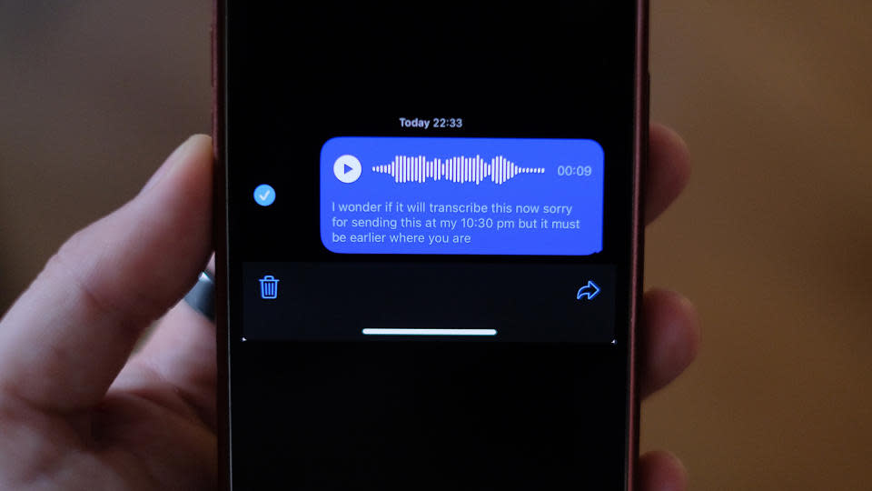 A person’s hand holding an iPhone. Its screen is mostly black (dark mode) with a blue box showing a transcription of an audio message. It reads, 