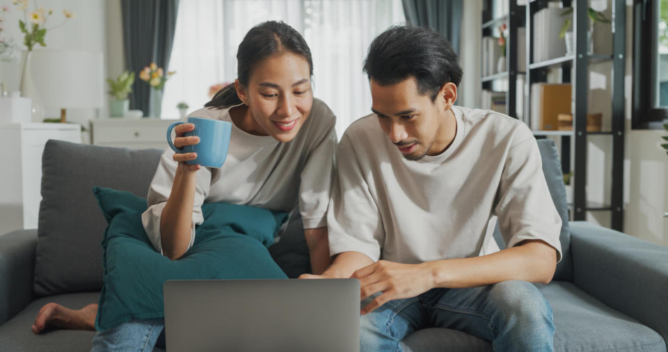 Happy young Asian couple sit on couch fun use laptop computer online shopping at home on holiday. Smiling young husband and wife laugh relax at home browsing application on gadgets, Lifestyle concept.