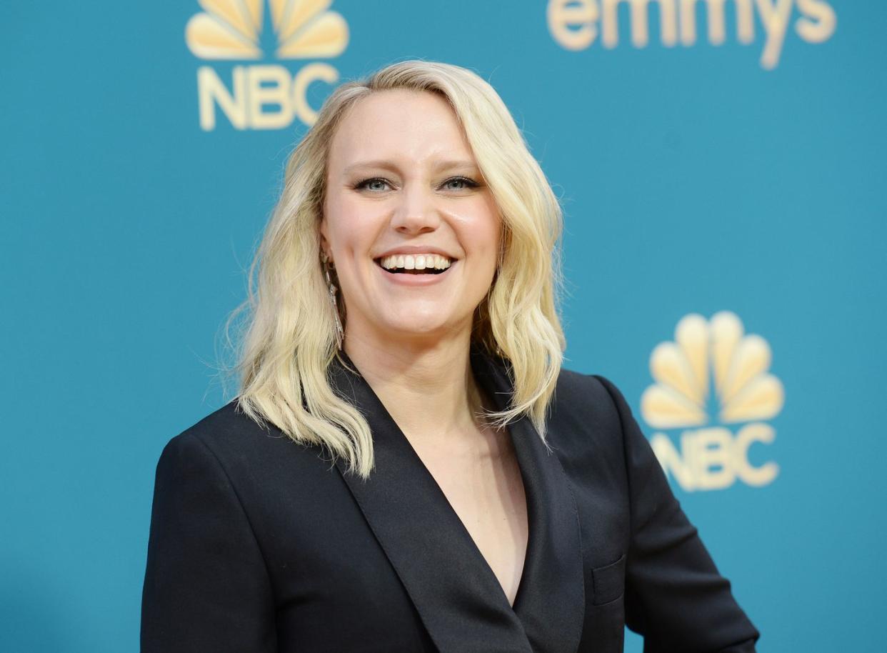 kate mckinnon, a woman stands smiling at the camera, blonde hair worn in loose waves to shoulders, wearing a black blazer
