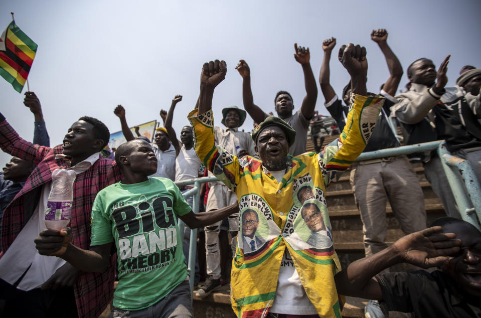 People in the stands sing songs, one wearing a jacket with the face of former president Robert Mugabe and another waving the national flag, as members of the public queue up to view his body at the Rufaro stadium in the capital Harare, Zimbabwe Friday, Sept. 13, 2019. The ongoing uncertainty of the burial of Mugabe, who died last week in Singapore at the age of 95, has eclipsed the elaborate plans for Zimbabweans to pay their respects to the former guerrilla leader at several historic sites. (AP Photo/Ben Curtis)