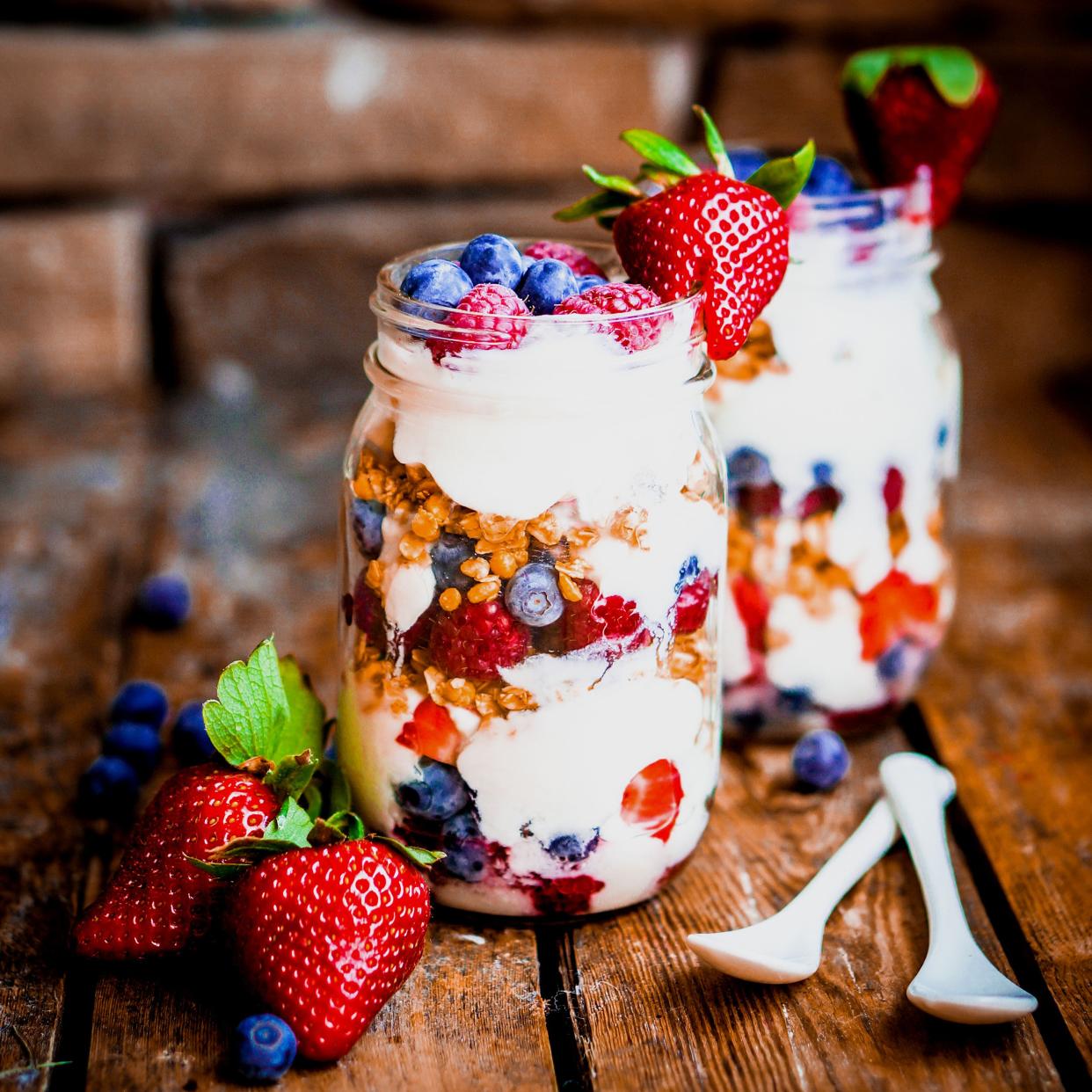 Two yogurt parfaits in mason glass jars with two porcelain spoons and surrounded by strawberries and blueberries, on a rustic wooden table with a blurred background of the table and a brick wall