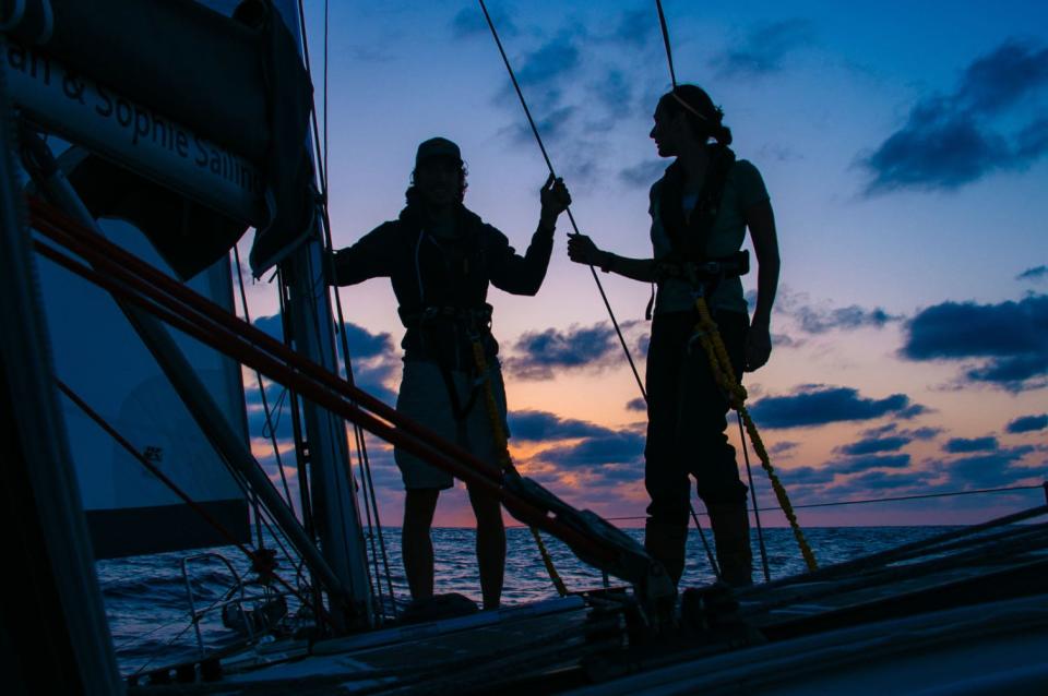 A couple stood on the deck of a sailboat in front of a sunset.