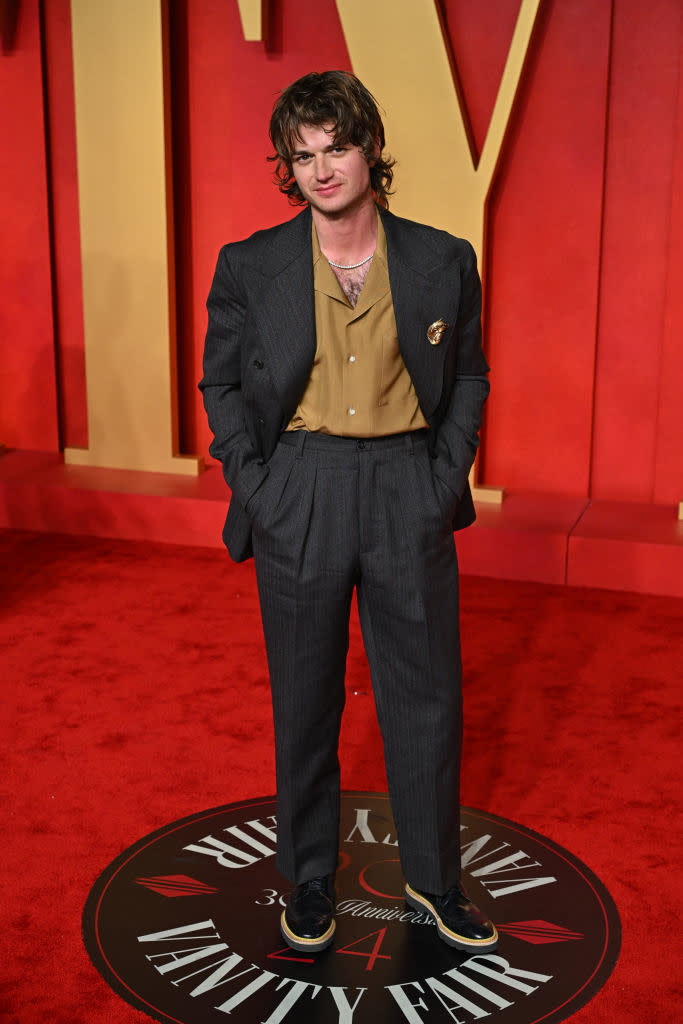 Man in pinstripe suit and gold shirt stands with hands in pockets on a red carpet event