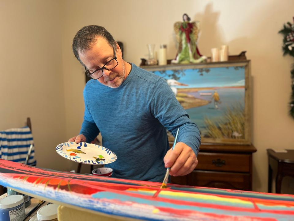 Keith White paints a mermaid scene on a surfboard at his home in Shallotte on Thursday, Dec. 14, 2023.