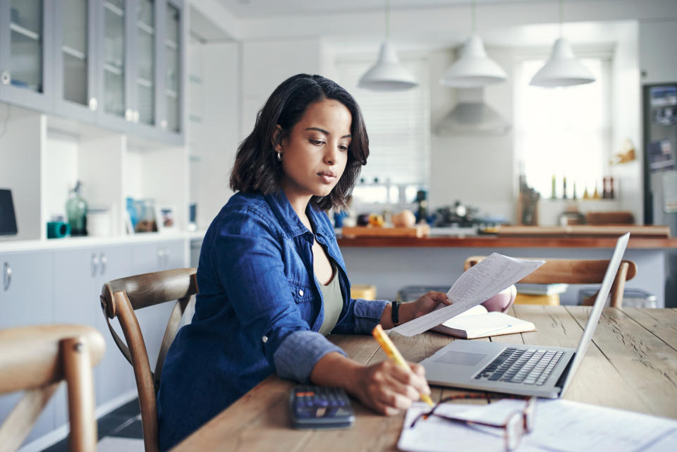 A woman holding papers and writing stuff down in front of her laptop