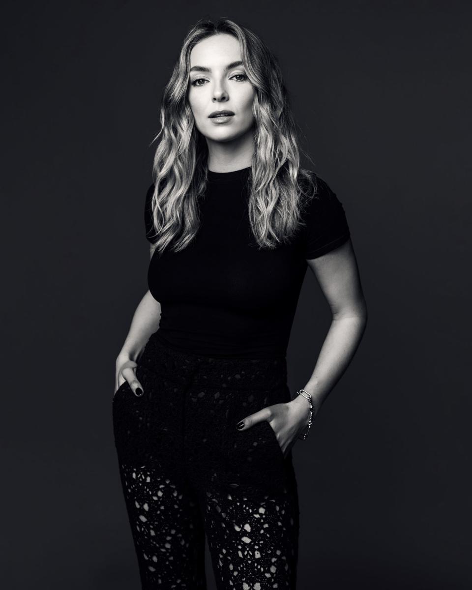 Jodie Comer poses in a stylish black outfit, featuring a t-shirt and patterned pants, with her hands in her pockets