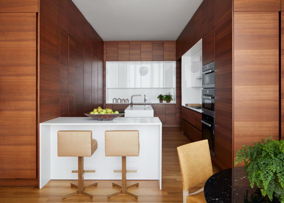 Warm, horizontal-grain walnut panels conceal storage cabinets and appliances in the kitchen. Scott created contrast and the illusion of a box within a box by covering the inset countertops and backsplash in white glass quartz. Fumed white oak and brass Walcott stools from KGBL provide counter seating.