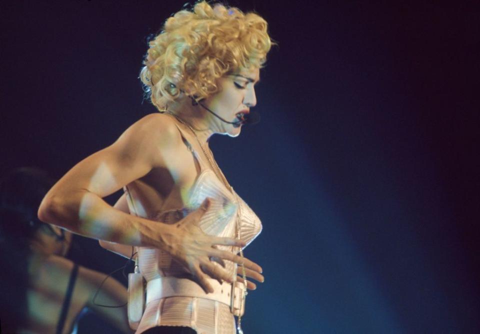 madonna, wearing a white outfit with her famous cone bra, singing into a headset microphone on a stage