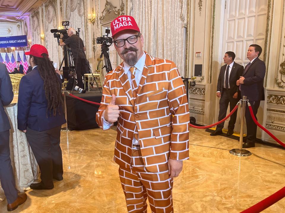 A man in a suit covered in brown bricks and a red hat that says "ultra-MAGA" gives a thumbs up sign