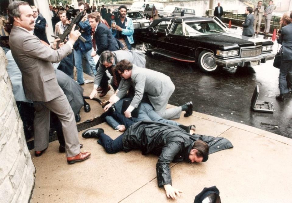 Several men in a chaotic scene with one lying on the ground, others standing with concerned expressions. A car is in the background