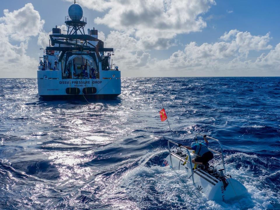 A submarine floats near the surface of the Pacific Ocean while a large research vessel is in the distance.