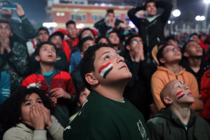 Children with Egyptian flags painted on their cheeks gaze up at a screen.