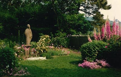 A statue of Virgin Mary stands in the center of a beautiful garden with colorful flowers, trees and plants.