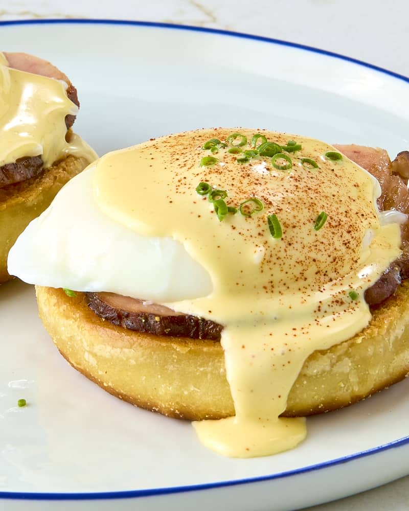 angled shot of eggs Benedict on a white plate with blue trim, topped with hollandaise sauce and chives