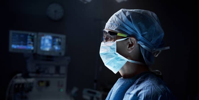 Person in surgical scrubs, mask, and cap stands in an operating room with medical monitors in the background