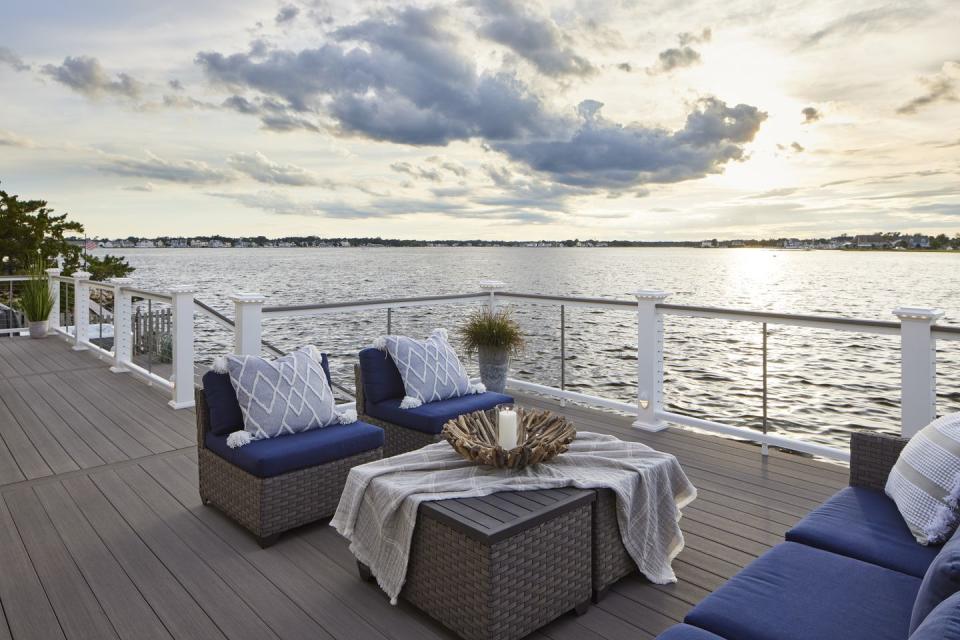 a composite deck is built overlooking the waters of a bay the sun is setting, and the sky features light pastel colors of orange and blue a large woven couch and two matching chairs sit around a coffee table the furniture features deep blue cushions and light blue throw pillows for a coastal feel a white composite railing with stainless steel cable rail infill offers clear sightlines to the water the gray composite decking is coastline from the timbertech azek vintage collection the railing is the classic composite series drink rail in white with a coastline deck board top rail