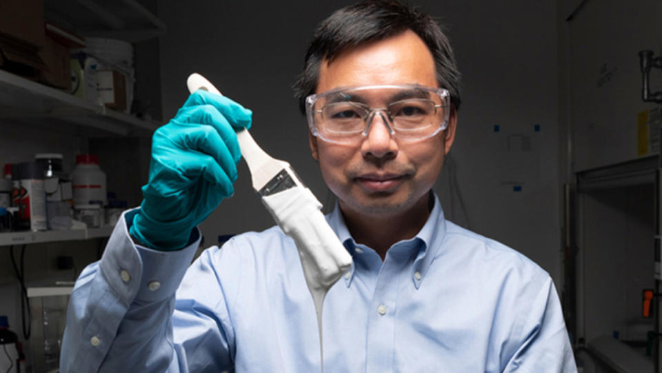 A scientist wearing clear safety glasses holds up a paintbrush dripping with an ultra-white paint.