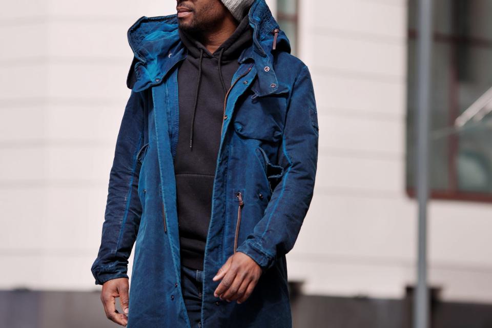 A man wears a blue jacket over a black hoodie while walking in the street past a beige building.