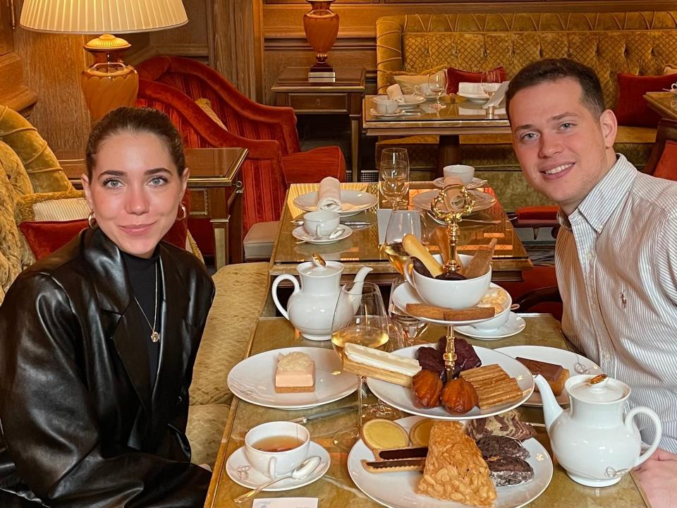 Dasha and her boyfriend sit at a table set up for tea. There is a three-tiered plate with pastries and two teapots.