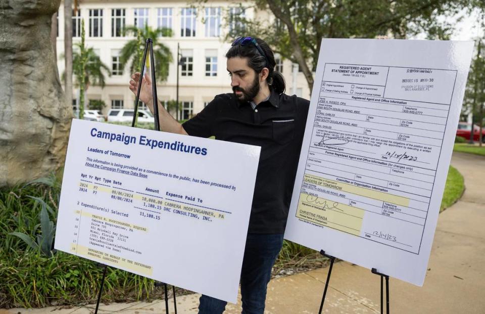 Kevin Lopez holds signs as Irina Vilarino, co-owner of Las Vegas Cuban Cuisine restaurant and candidate for Doral City Council Seat 3, holds a press conference at Downtown Doral Park on Wednesday, August 28, 2024. Public documents are displayed showing Mayor Christi Fraga's involvement in a complaint challenging her candidacy for council member.