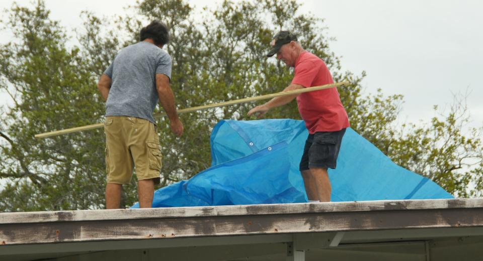 Two men install a blue tarp atop a storm-damaged roof Monday morning in the River Grove Mobile Home Village in Micco.