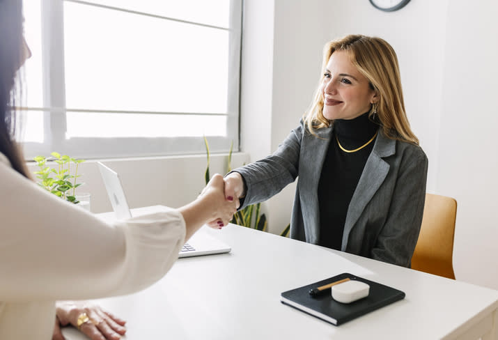 A woman in a grey blazer and black turtleneck smiles while shaking hands with another person at a desk in an office