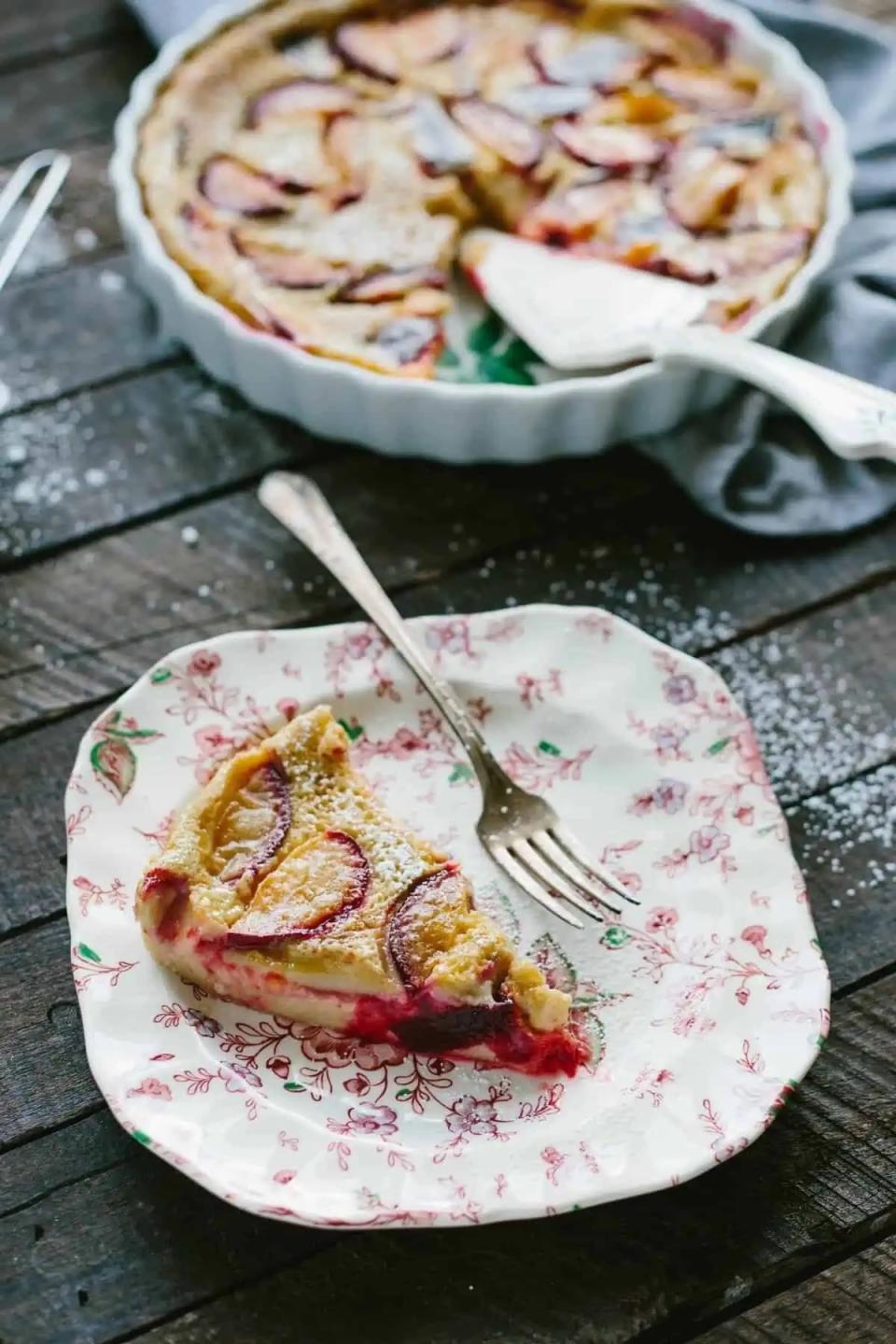 A slice of fruit tart on a decorative plate with a fork, and a larger tart with a missing slice in the background on a wooden table