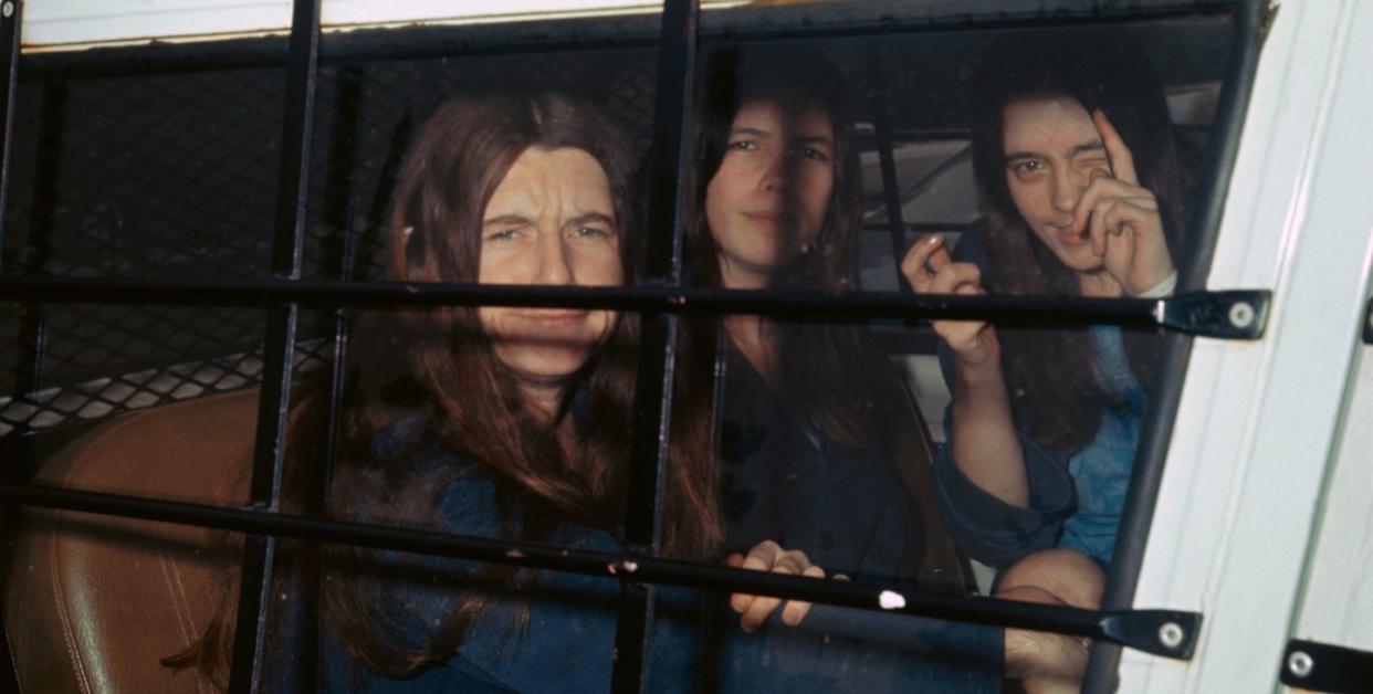 three female members of the manson family sit inside a police vehicle with cars on the window