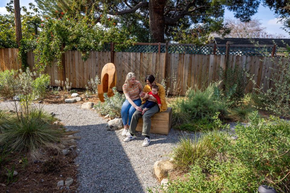 Vince Skelly holds his son with his wife sitting beside him on a wood block in his yard.