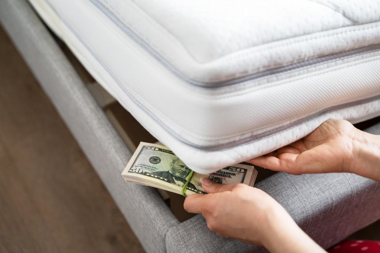 two hands placing a stack of money under the corner of a mattress