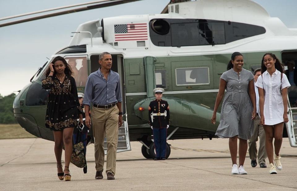 Barack Obama in casual shirt and slacks walks with Michelle in a dress and their daughters, near a helicopter, with a marine saluting