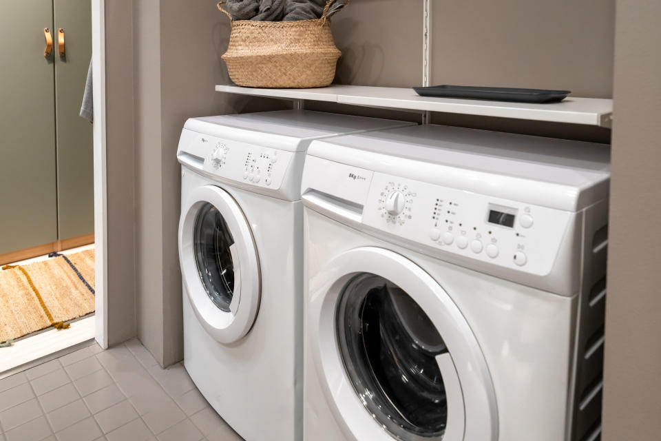 Two modern front-load washing machines are situated side by side in a laundry room. A basket of folded laundry is on a shelf above them