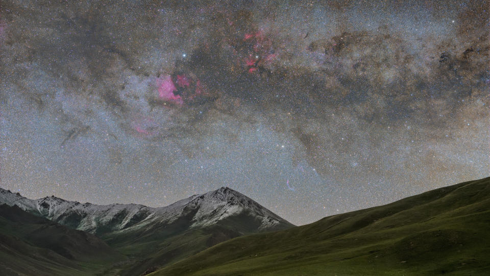     The night sky is full of stars with snow-capped mountains and a huge emission nebula glowing pink-red. 