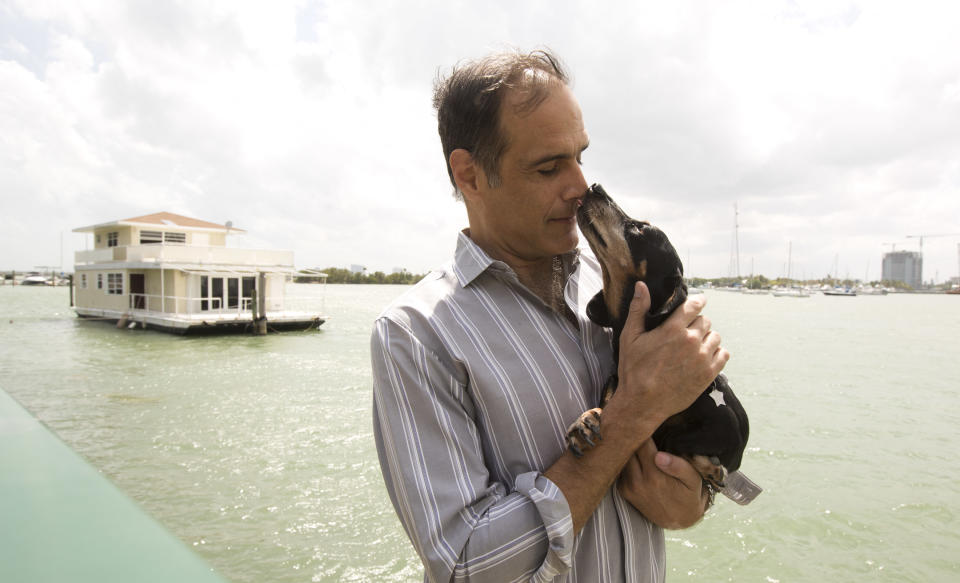 In this April 22, 2014 photo, Fane Lozman poses for photos holding his dog and in front of his home floating in the waters near North Bay Village, Fla. He caught legal lightning in a bottle last year when the U.S. Supreme Court agreed with him that his floating home was a house, not a vessel covered by maritime law. But the justices haven’t had the last word: Lozman is still fighting for compensation for the home, which was destroyed years ago. The Fort Lauderdale-based federal judge whose decision on the floating home was overturned, U.S. District Judge William Dimitrouleas, refused earlier this year to give Lozman any of the $25,000 bond posted by the city of Riviera Beach to pay for Lozman’s home in case he won. (AP Photo/J Pat Carter)