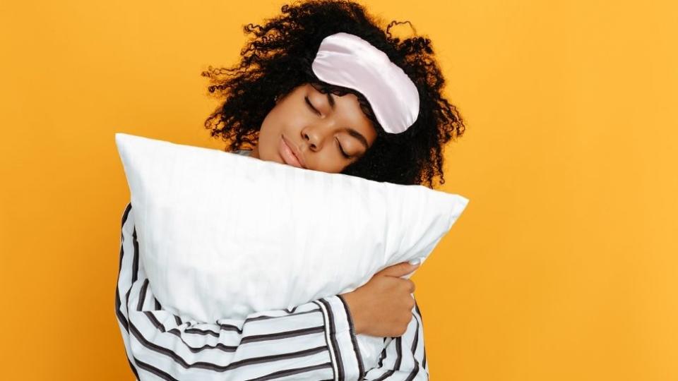 A woman in pyjamas with tight curly hair cuddles a pillow and has a sleep mask ready to be worn.