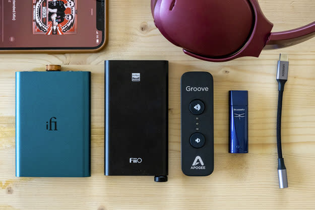 Image of a series of portable DACs on a wooden table, as well as an iPhone screen and a pair of headphones.