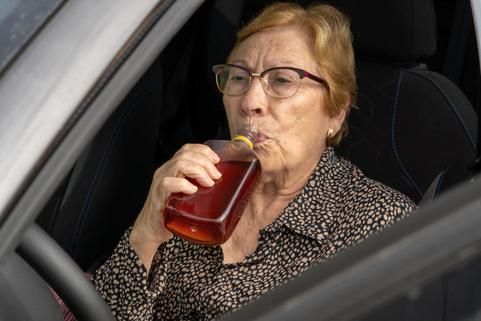 An elderly woman in glasses and a patterned blouse drinks from a bottle while sitting in a car