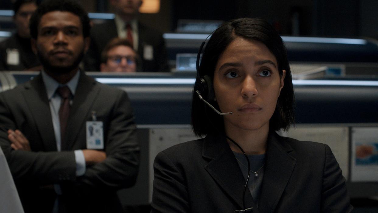  Woman with shoulder length black hair wearing a suit and a headset. She is in a control center with lots of other people behind her also wearing suits and special badges. 