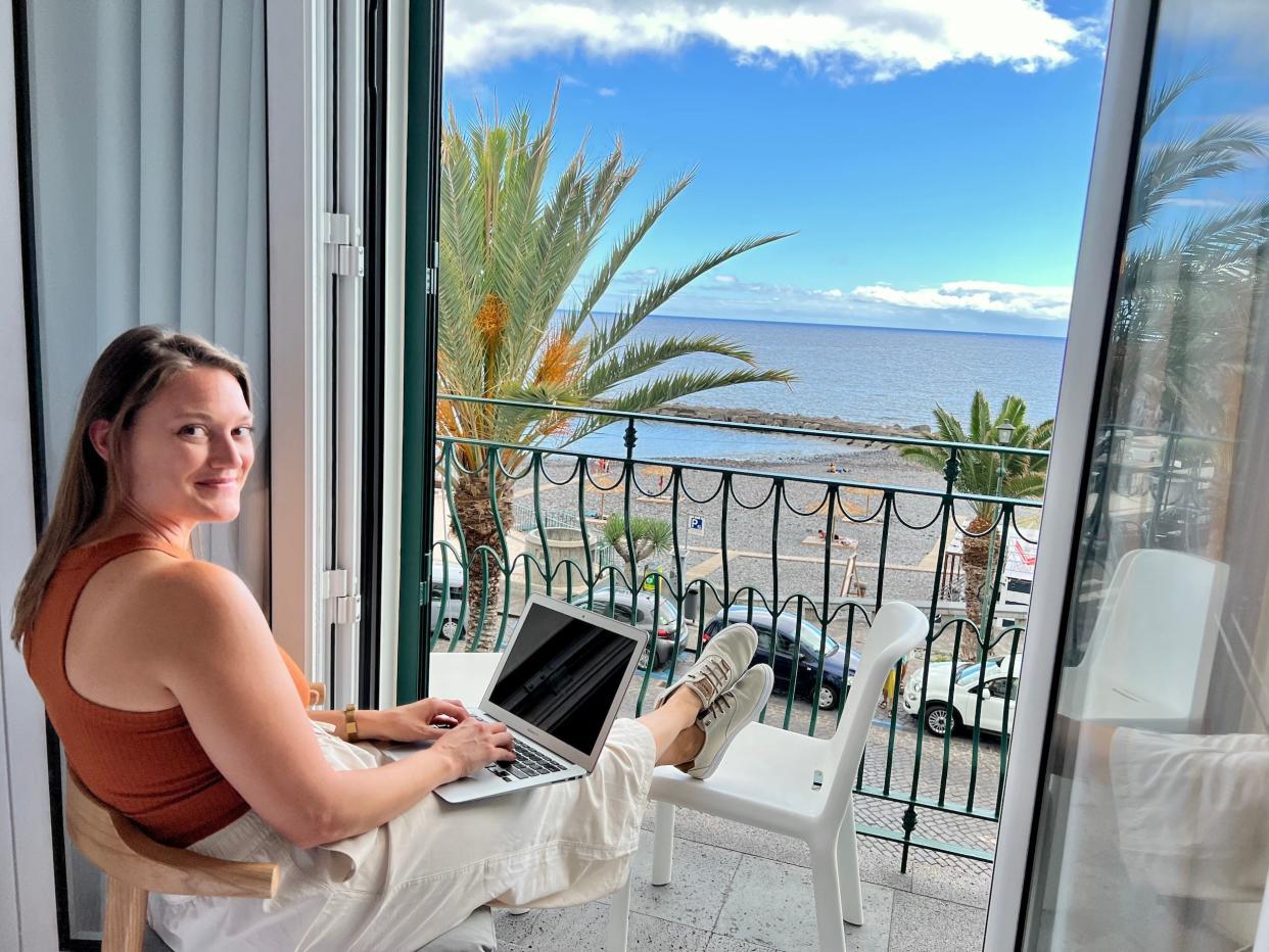 A woman sits on a balcony overlooking the sea with a laptop in her lap. She smiles at the camera.