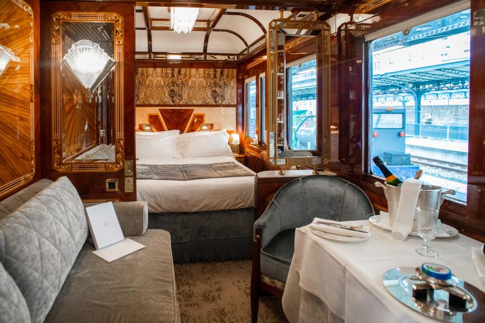 Inside a wood-walled train suite with a seat on the right a couch on the left, and a bed in the back center.