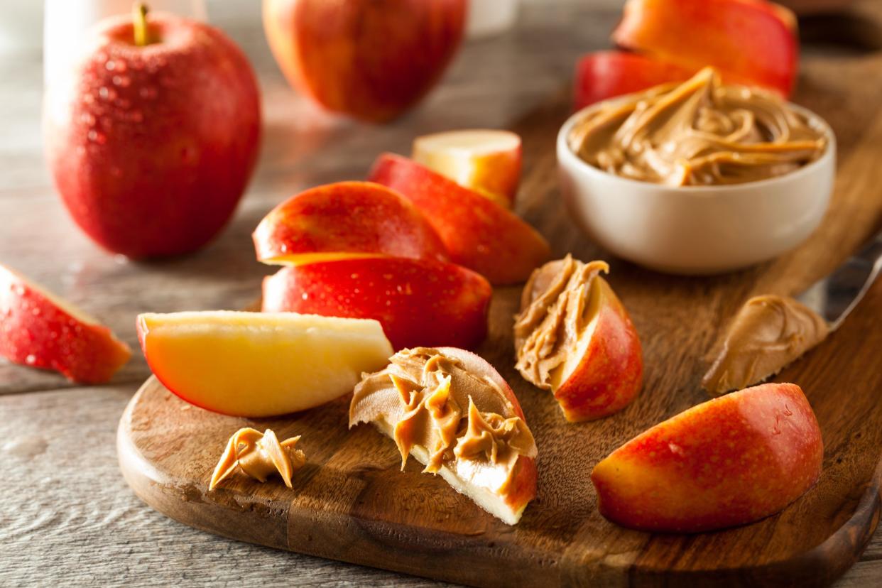 Slices of red apples, some with peanut butter with a knife with peanut butter and a small white bowl with peanut butter, all on a wooden serving board, surrounded by whole apples and slices of apples on a wooden table