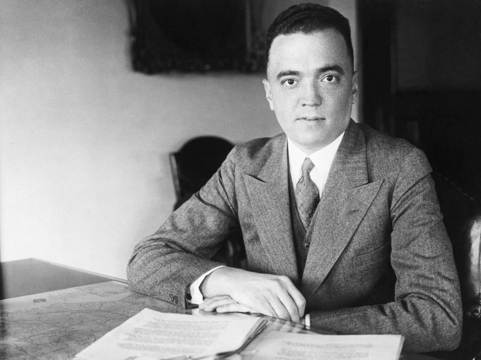 J. Edgar Hoover sits at a desk with a stack of papers before him.