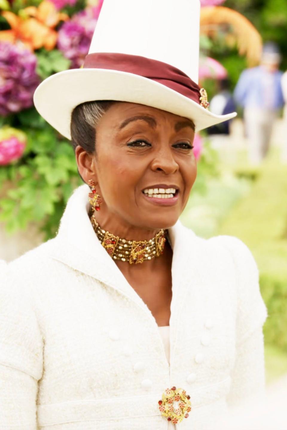 Adjoa Andoh as Lady Danbury at an outdoor formal event wearing a suit jacket, gold jewelry, and a matching top hat. Colorful flowers in the background