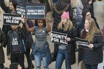 Congregated protesters carry signs that read "shut down ableism" and "disability rights are human rights" and "dignity for all"