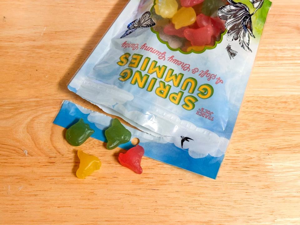 A bag of gummy candies with four candies spilling out onto a wooden table. The bag has illustrations of grass and a sky on it