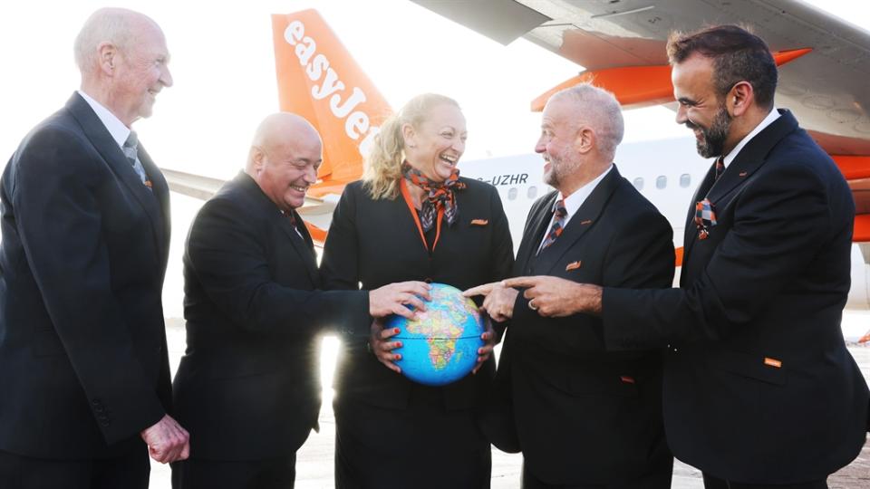 Older easyJet cabin crew staff stand beside one of the airline's planes. One holds a globe as they all share a joke.