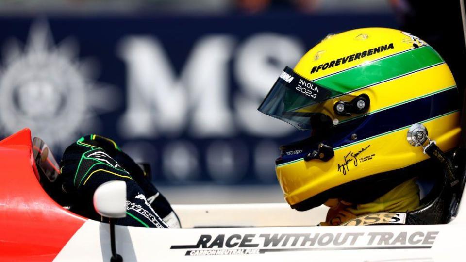 A close-up side image of Sebastian Vettel driving Ayrton senna's 1993 McLaren while wearing a helmet with the Senna yellow, green and blue design