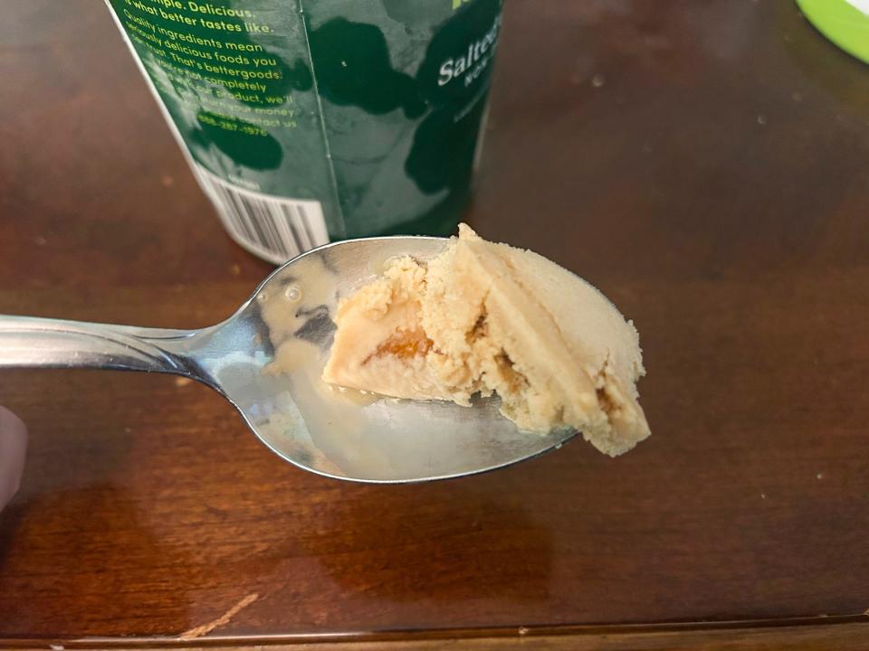 A metal spoon with a scoop of tan ice cream with a caramel swirl in it and a green carton in the background