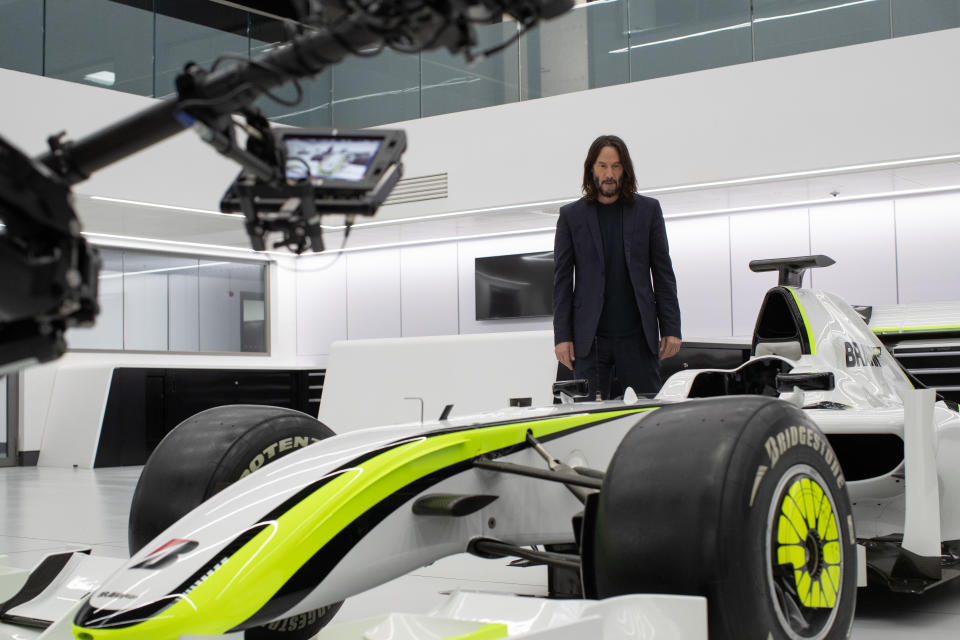 Keanu Reeves is dressed in black, standing behind a white and neon green Formula One car. (Disney+)